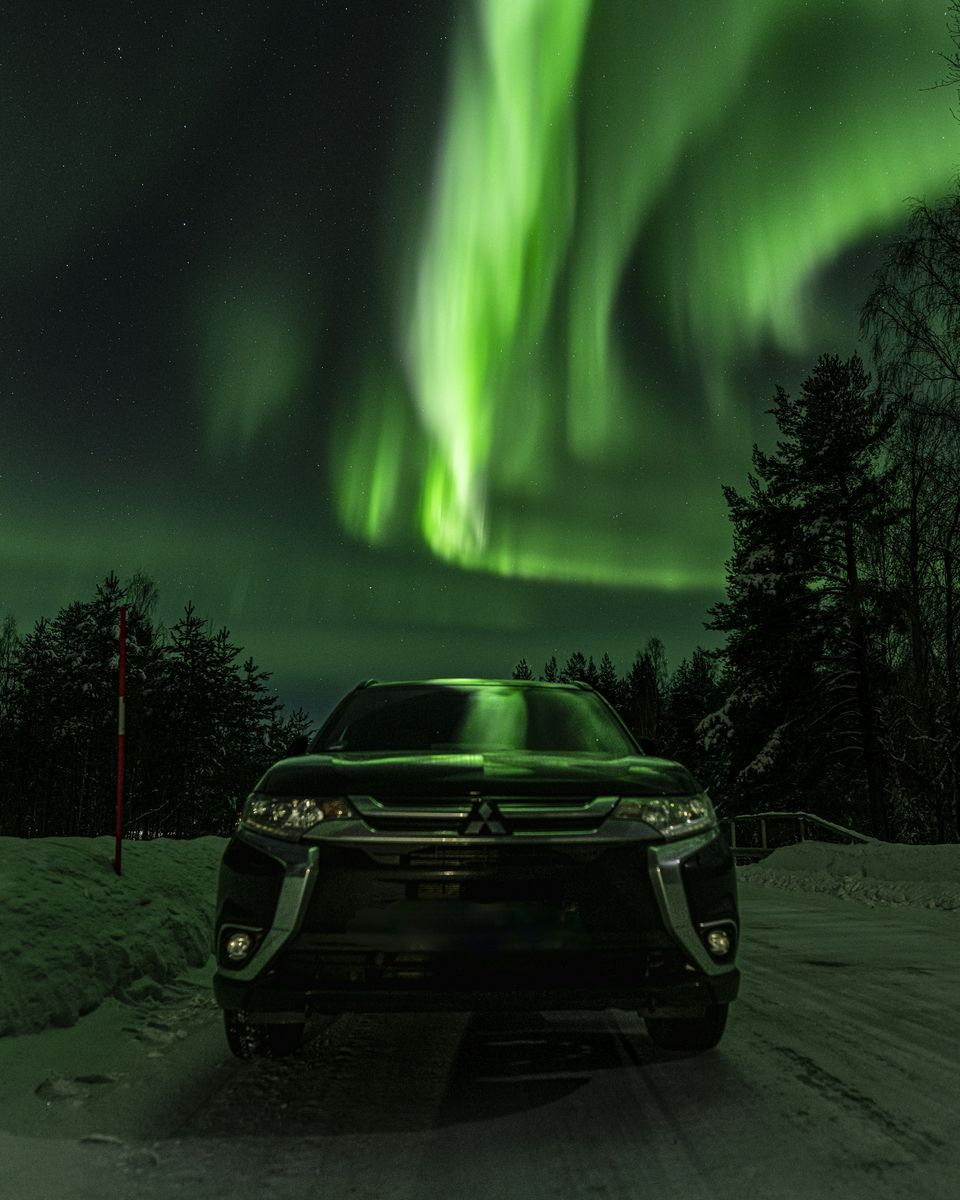 An SUV with its lights on under a bright green aurora in a snowy forest