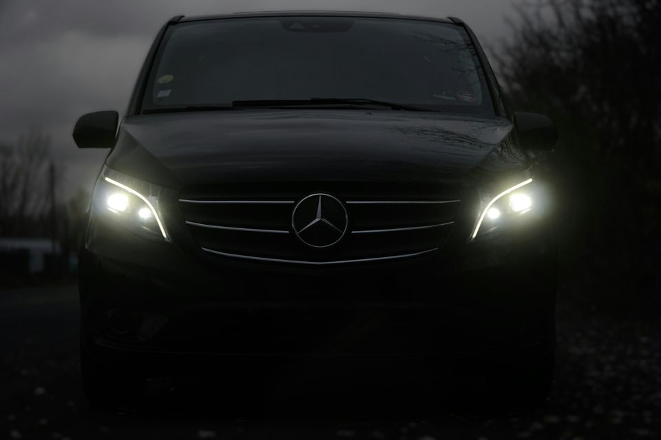The front of a dark Mercedes-Benz van with its headlights on